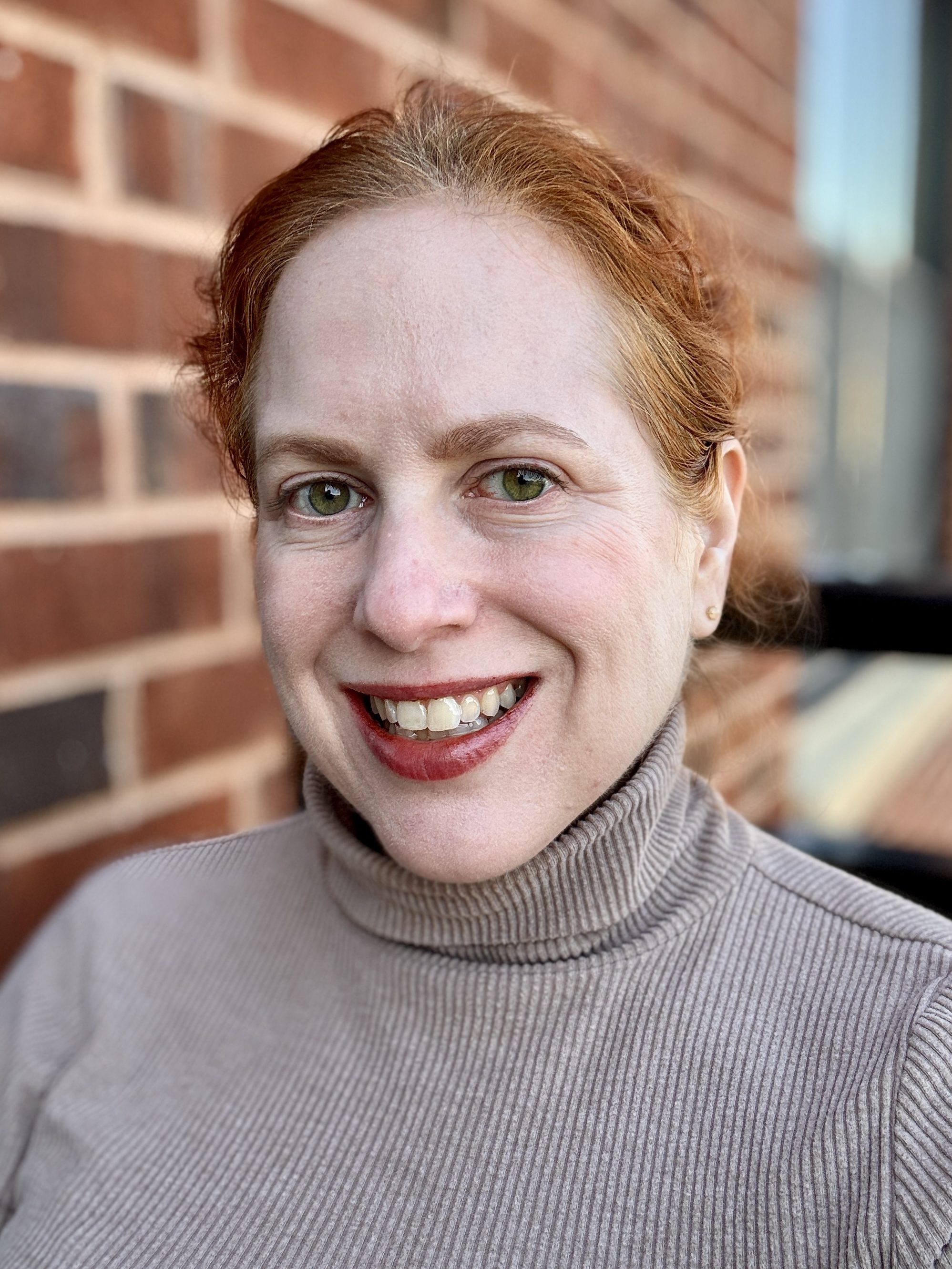 A red-haired woman wearing a turtleneck sits in front of a brick wall.